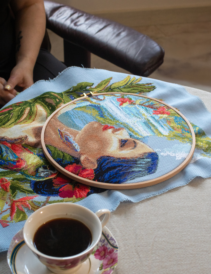 A completed cross-stitch of a woman with red hibiscus flowers in her hair, a colorful parrot on her shoulder, tropical leaves in the foreground, and waterfalls in the background.