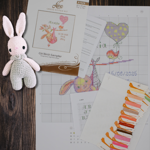 A completed cross-stitch birth sampler featuring a bunny in a pink sling hanging from a branch, with a bird holding a pink heart balloon and space for baby details.