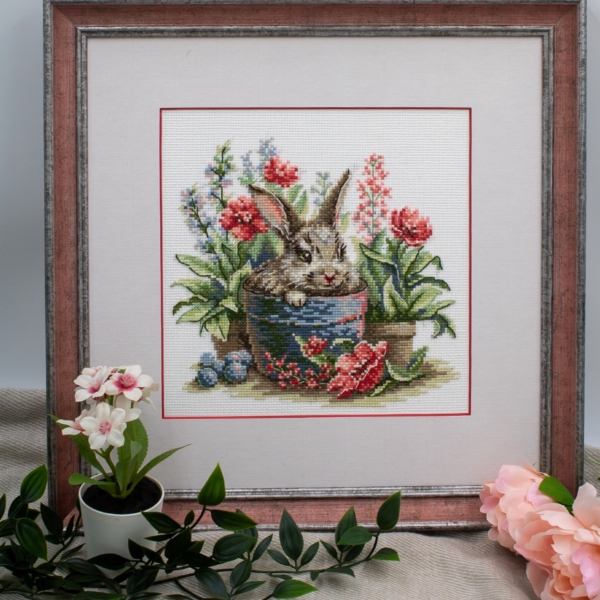 A completed cross-stitch of a brown bunny peeking out of a blue flower pot surrounded by red flowers and green plants in a garden scene.