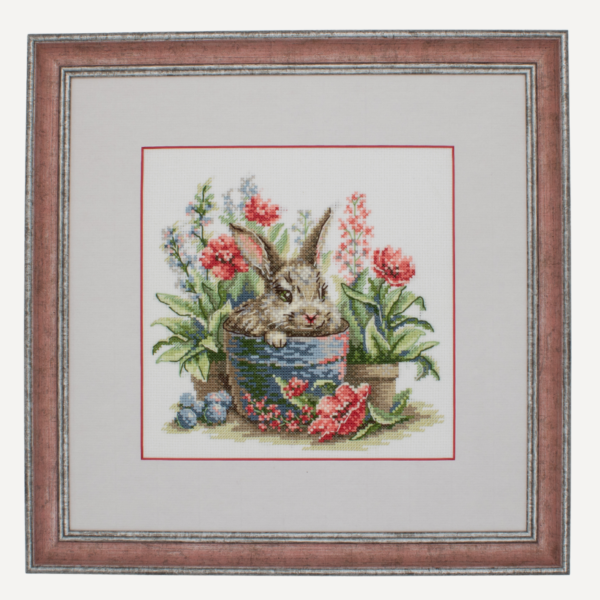 A completed cross-stitch of a brown bunny peeking out of a blue flower pot surrounded by red flowers and green plants in a garden scene.