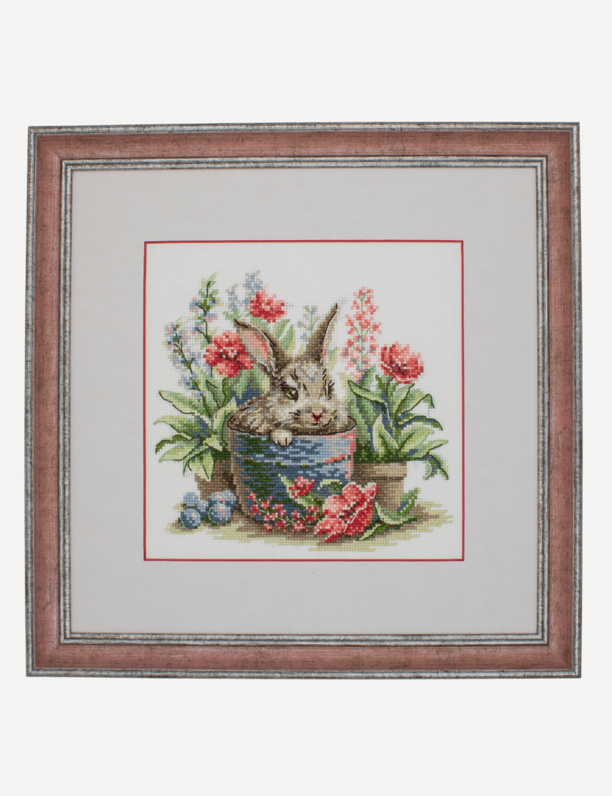 A completed cross-stitch of a brown bunny peeking out of a blue flower pot surrounded by red flowers and green plants in a garden scene.