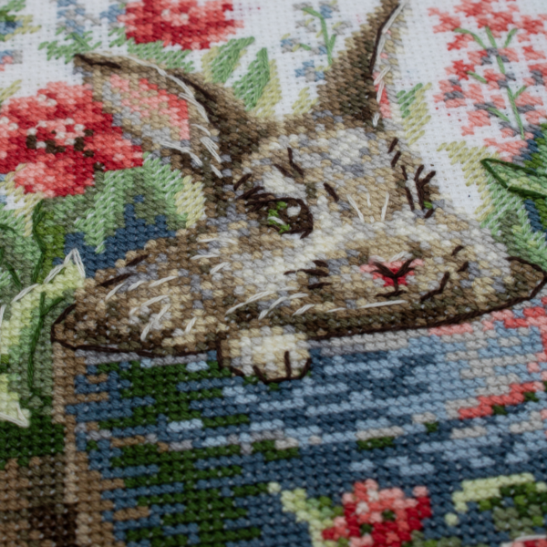 A completed cross-stitch of a brown bunny peeking out of a blue flower pot surrounded by red flowers and green plants in a garden scene.