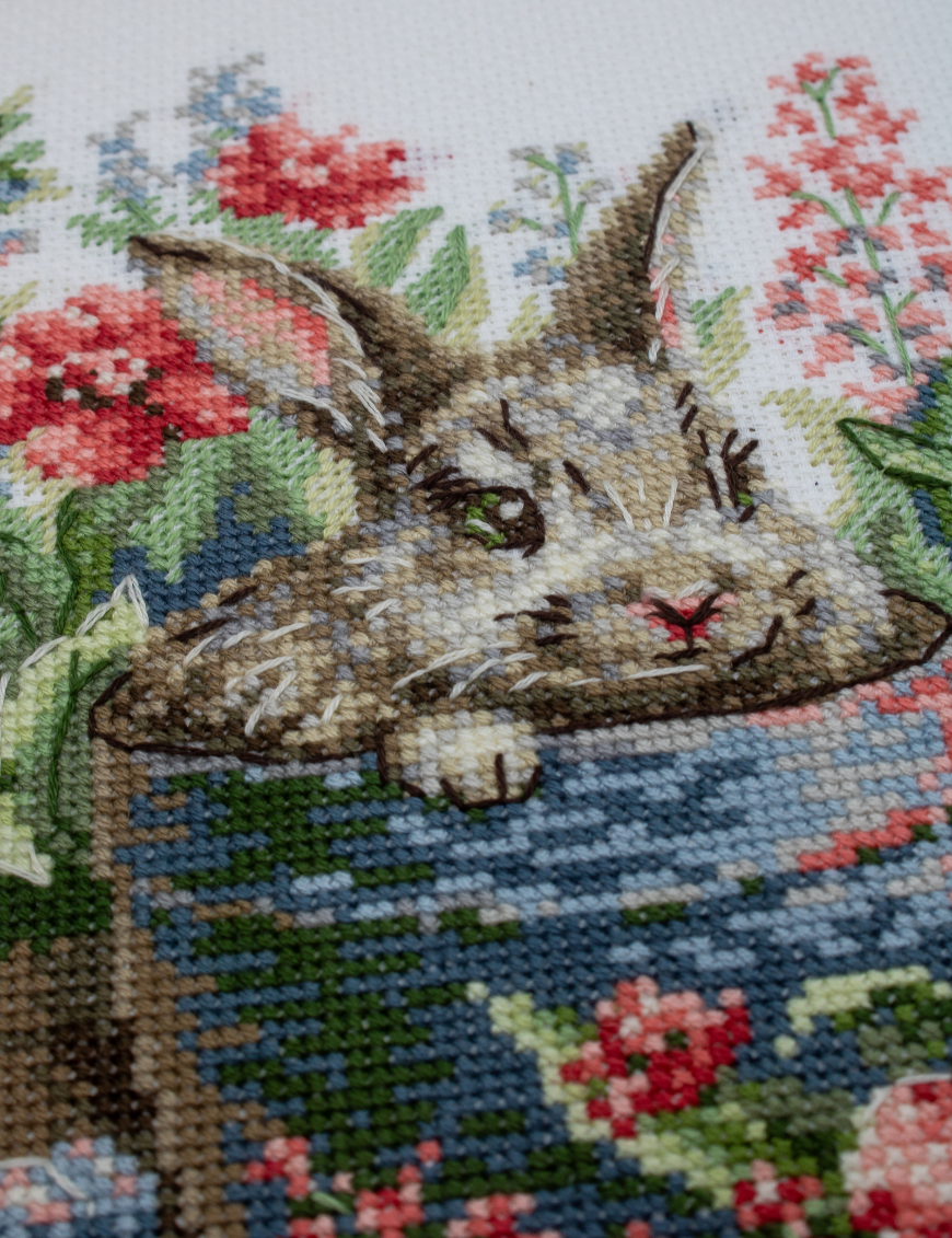 A completed cross-stitch of a brown bunny peeking out of a blue flower pot surrounded by red flowers and green plants in a garden scene.