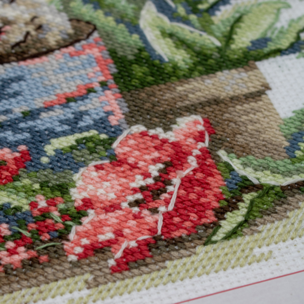A completed cross-stitch of a brown bunny peeking out of a blue flower pot surrounded by red flowers and green plants in a garden scene.