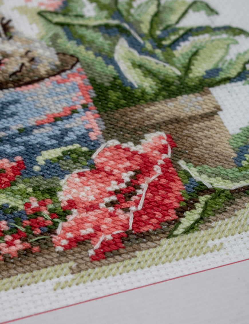 A completed cross-stitch of a brown bunny peeking out of a blue flower pot surrounded by red flowers and green plants in a garden scene.