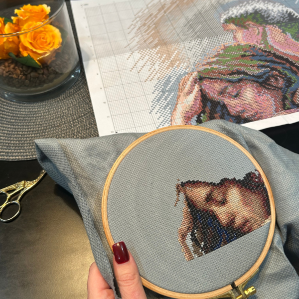 A completed cross-stitch of a couple embracing tenderly, shown in soft natural tones against a light blue background.