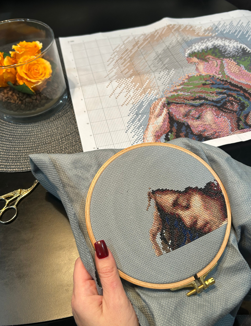 A completed cross-stitch of a couple embracing tenderly, shown in soft natural tones against a light blue background.