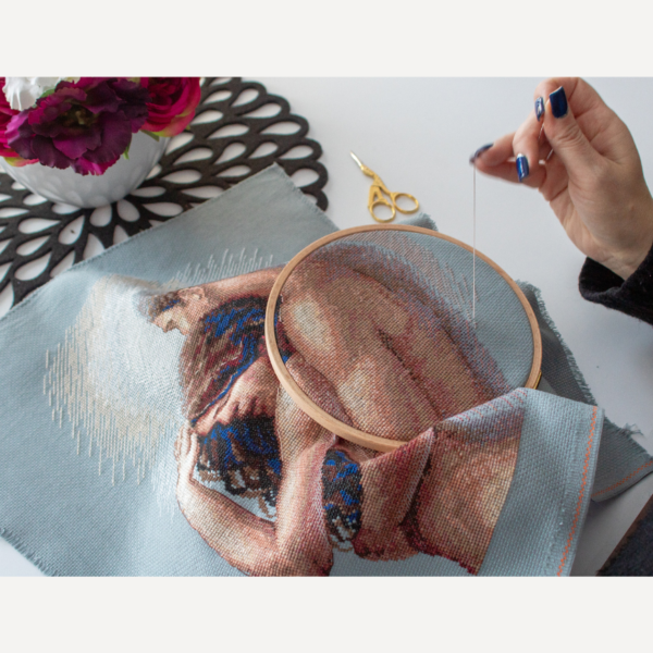 A completed cross-stitch of a couple embracing tenderly, shown in soft natural tones against a light blue background.