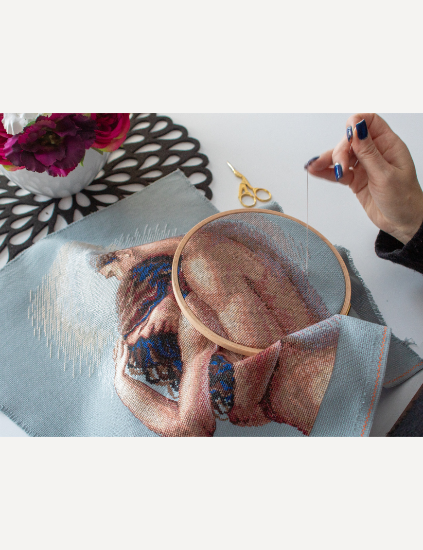 A completed cross-stitch of a couple embracing tenderly, shown in soft natural tones against a light blue background.