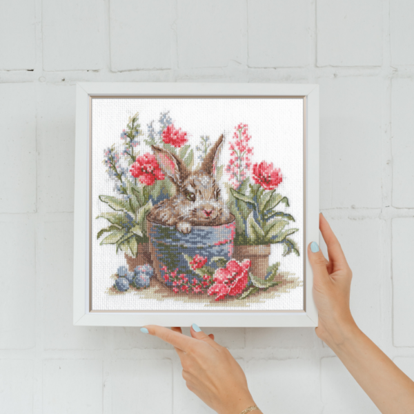 A completed cross-stitch of a brown bunny peeking out of a blue flower pot surrounded by red flowers and green plants in a garden scene.