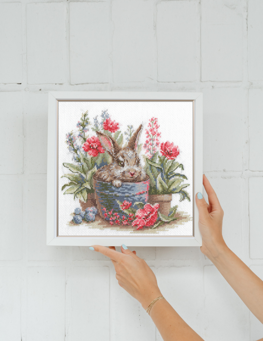 A completed cross-stitch of a brown bunny peeking out of a blue flower pot surrounded by red flowers and green plants in a garden scene.