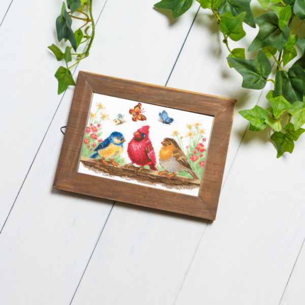 A completed cross-stitch of three colorful birds perched on a branch with butterflies and spring flowers in the background.