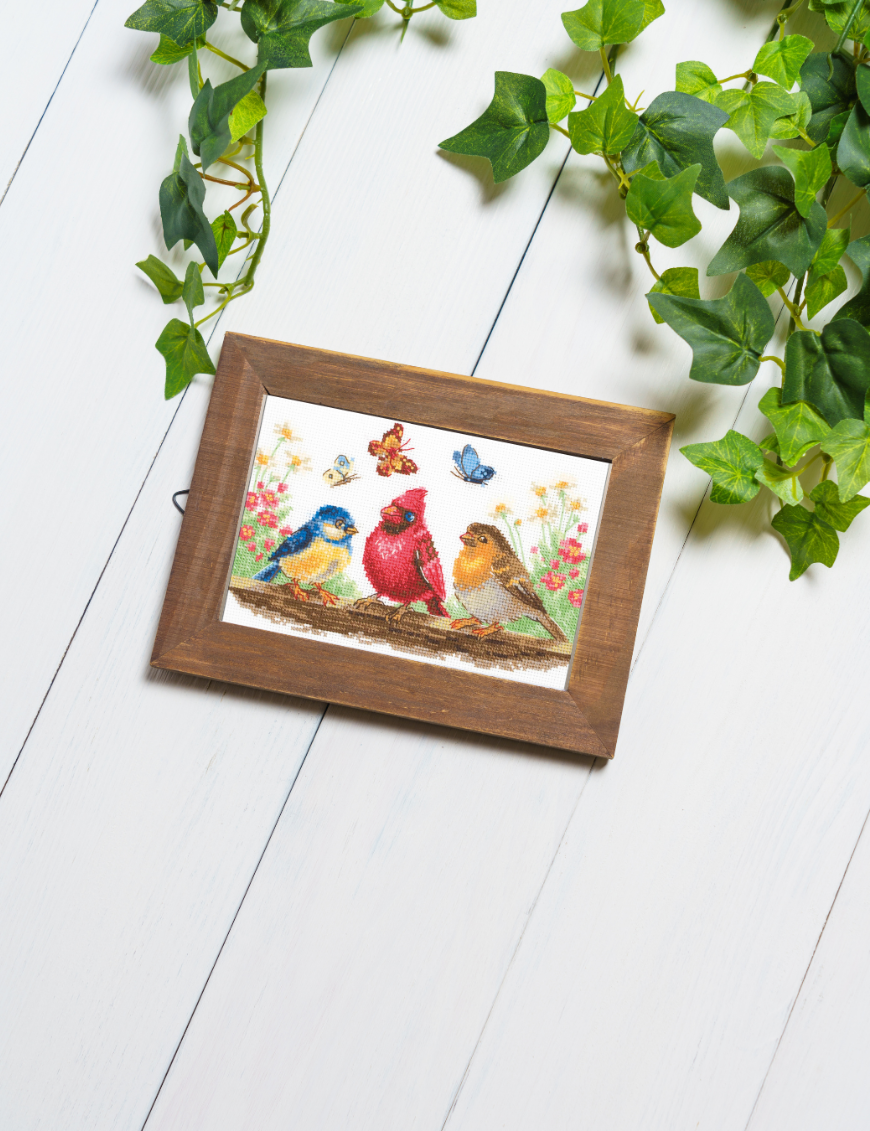 A completed cross-stitch of three colorful birds perched on a branch with butterflies and spring flowers in the background.