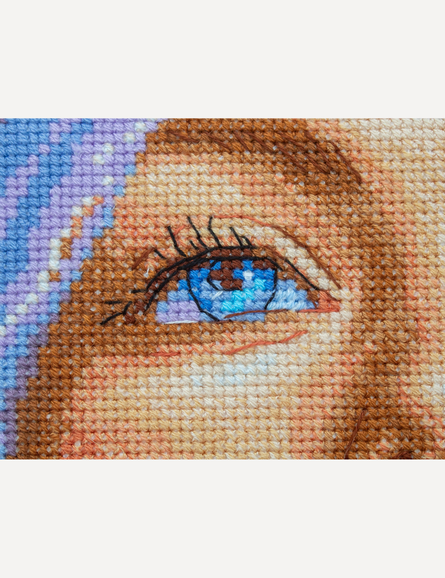 A completed cross-stitch of a girl with pastel purple hair blowing a pink bubblegum bubble, with bright blue eyes and soft blended tones.