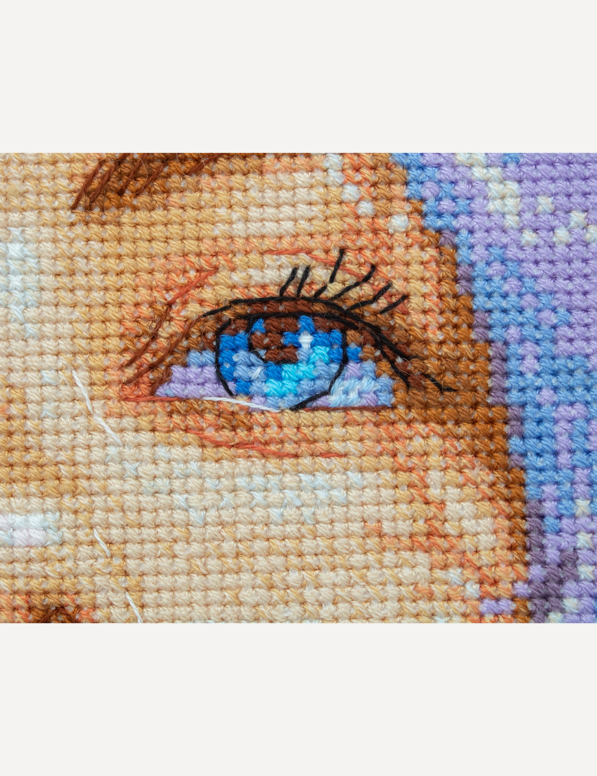 A completed cross-stitch of a girl with pastel purple hair blowing a pink bubblegum bubble, with bright blue eyes and soft blended tones.