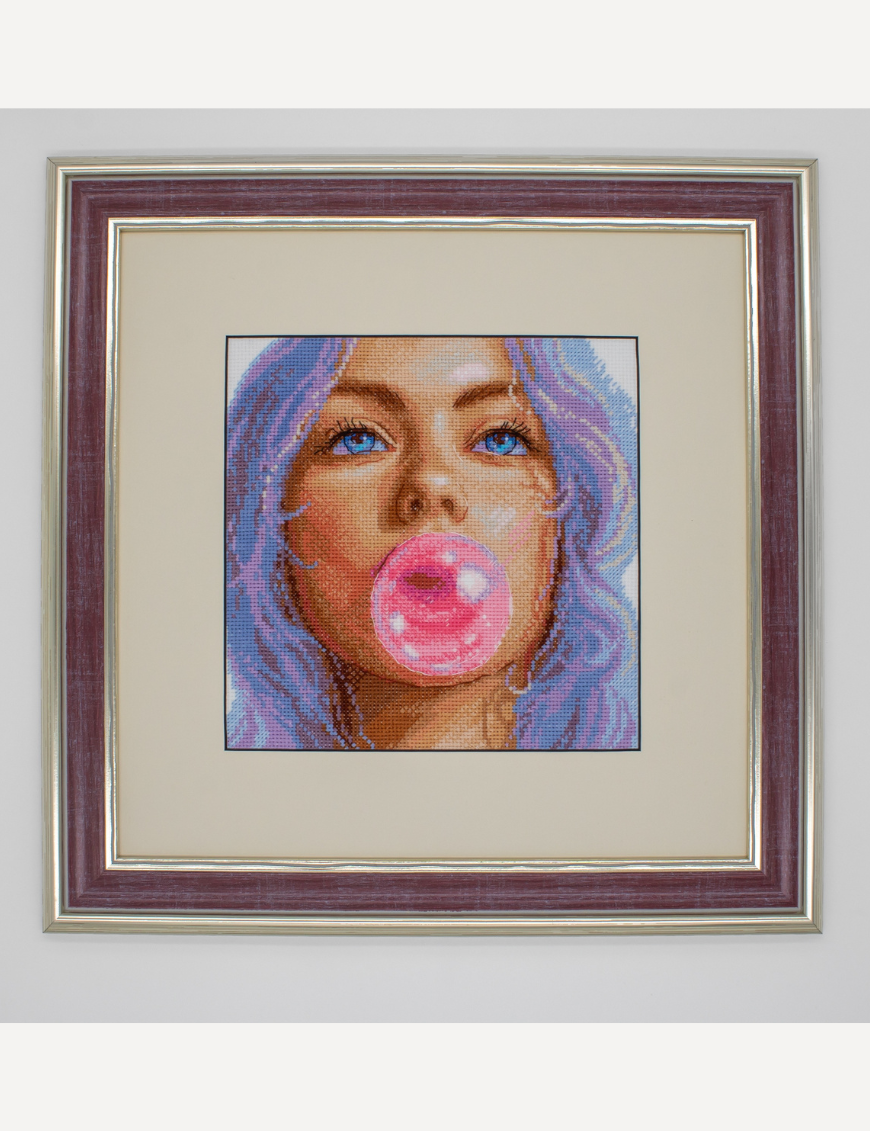 A completed cross-stitch of a girl with pastel purple hair blowing a pink bubblegum bubble, with bright blue eyes and soft blended tones.