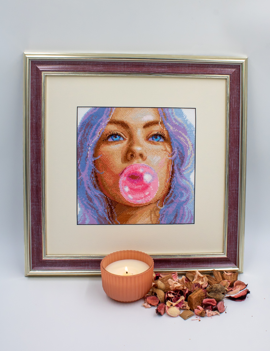 A completed cross-stitch of a girl with pastel purple hair blowing a pink bubblegum bubble, with bright blue eyes and soft blended tones.