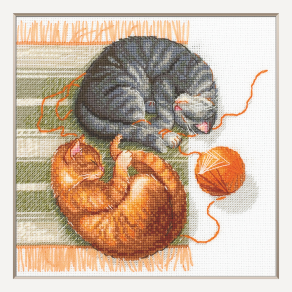 A completed cross-stitch of two cats resting on a rug, playing with orange yarn balls, stitched in warm brown, grey, and orange tones.