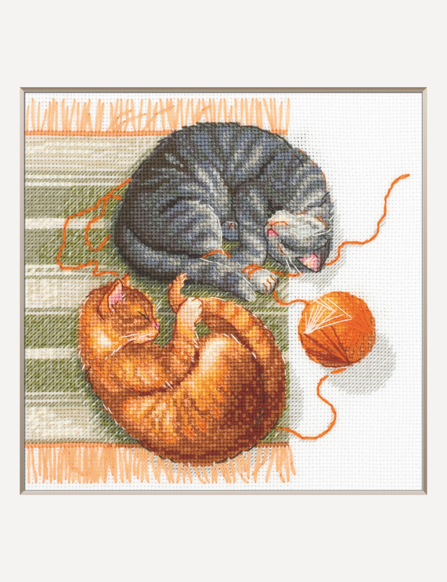 A completed cross-stitch of two cats resting on a rug, playing with orange yarn balls, stitched in warm brown, grey, and orange tones.