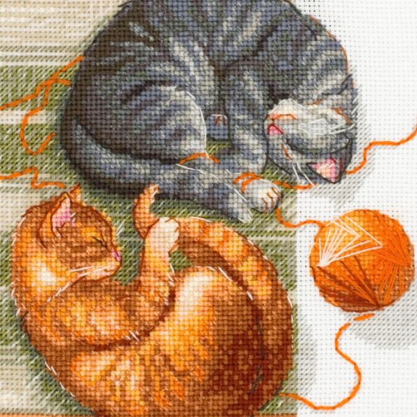 A completed cross-stitch of two cats resting on a rug, playing with orange yarn balls, stitched in warm brown, grey, and orange tones.