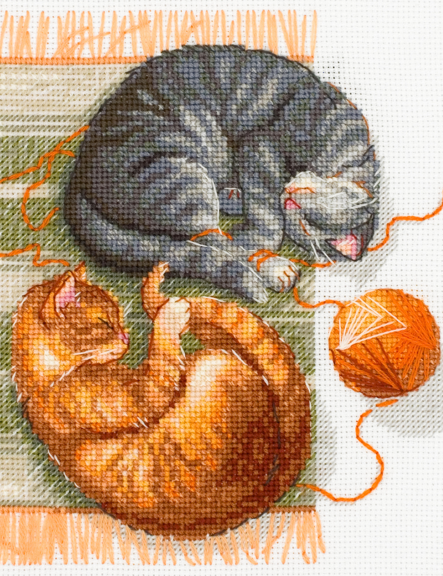 A completed cross-stitch of two cats resting on a rug, playing with orange yarn balls, stitched in warm brown, grey, and orange tones.