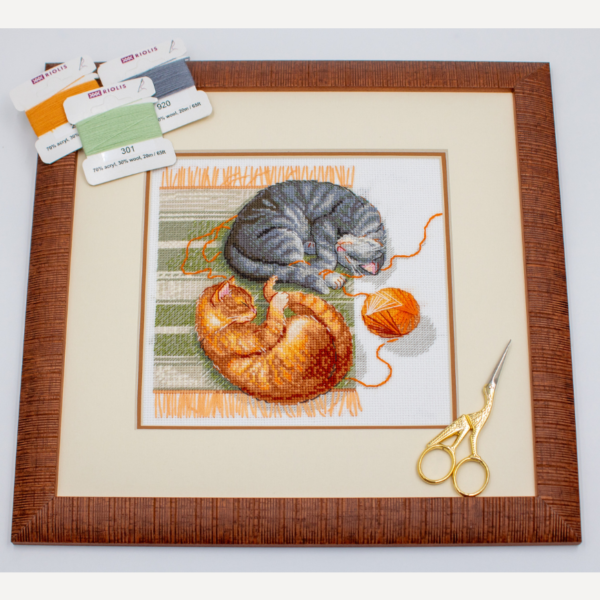 A completed cross-stitch of two cats resting on a rug, playing with orange yarn balls, stitched in warm brown, grey, and orange tones.