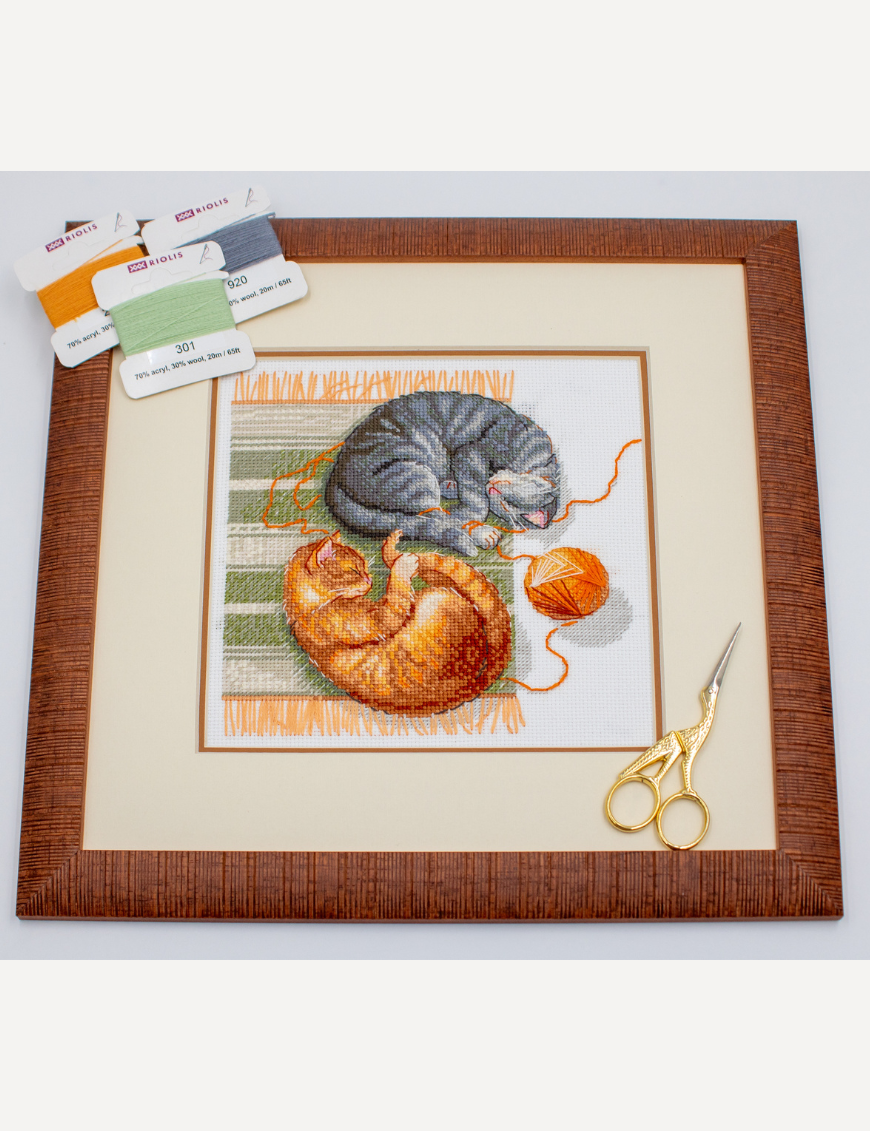 A completed cross-stitch of two cats resting on a rug, playing with orange yarn balls, stitched in warm brown, grey, and orange tones.