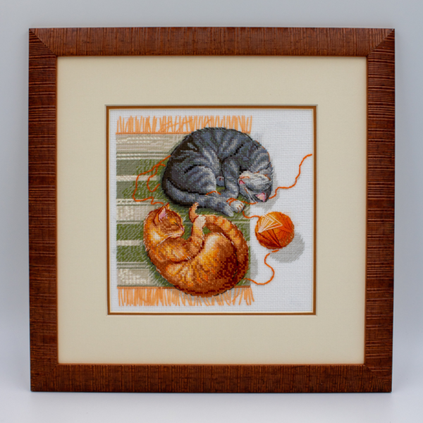 A completed cross-stitch of two cats resting on a rug, playing with orange yarn balls, stitched in warm brown, grey, and orange tones.