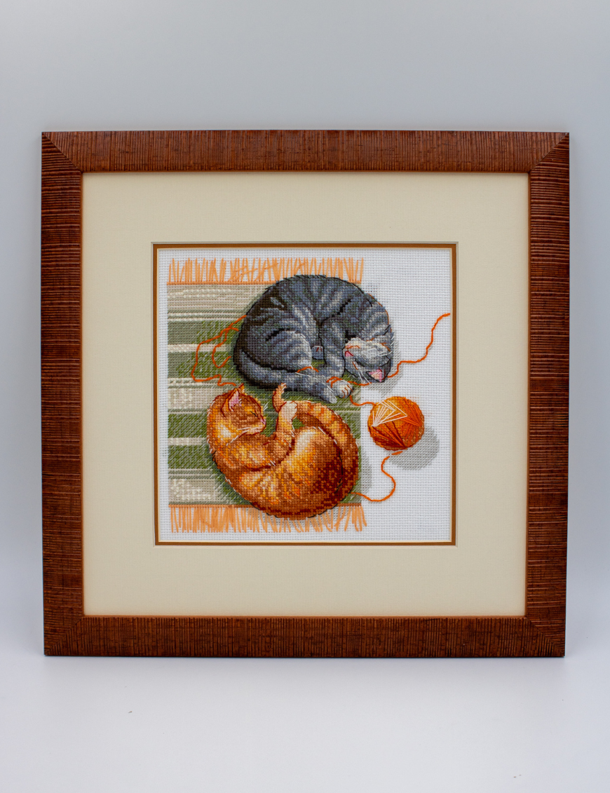 A completed cross-stitch of two cats resting on a rug, playing with orange yarn balls, stitched in warm brown, grey, and orange tones.
