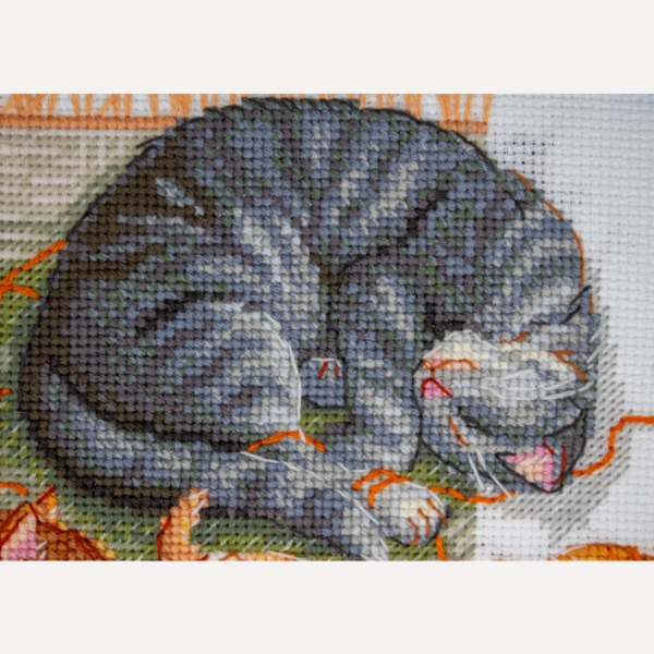 A completed cross-stitch of two cats resting on a rug, playing with orange yarn balls, stitched in warm brown, grey, and orange tones.