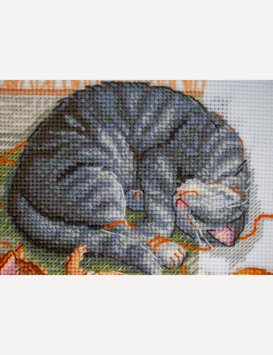 A completed cross-stitch of two cats resting on a rug, playing with orange yarn balls, stitched in warm brown, grey, and orange tones.
