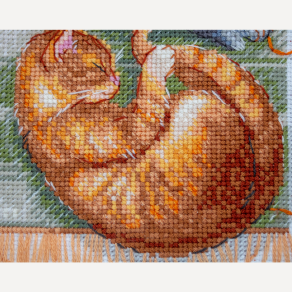 A completed cross-stitch of two cats resting on a rug, playing with orange yarn balls, stitched in warm brown, grey, and orange tones.