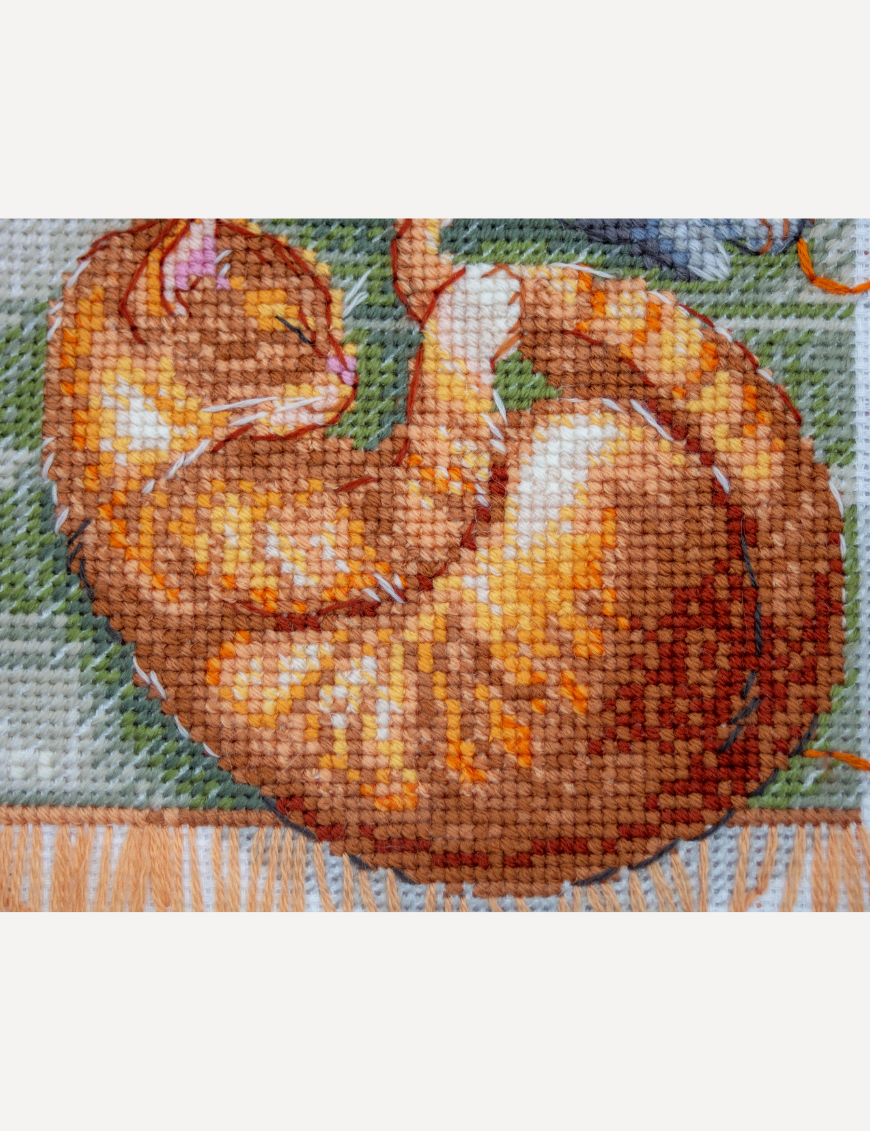 A completed cross-stitch of two cats resting on a rug, playing with orange yarn balls, stitched in warm brown, grey, and orange tones.