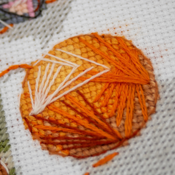 A completed cross-stitch of two cats resting on a rug, playing with orange yarn balls, stitched in warm brown, grey, and orange tones.