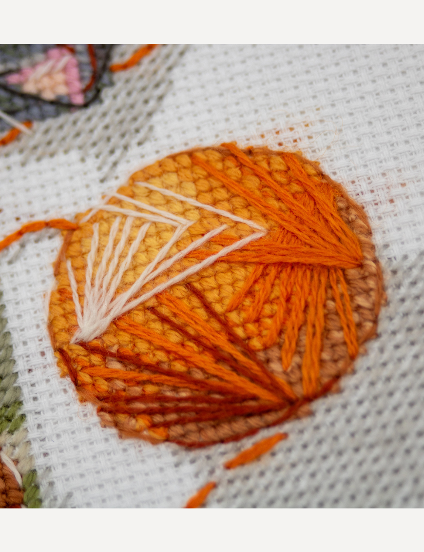 A completed cross-stitch of two cats resting on a rug, playing with orange yarn balls, stitched in warm brown, grey, and orange tones.