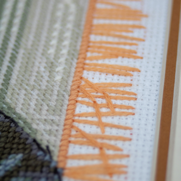 A completed cross-stitch of two cats resting on a rug, playing with orange yarn balls, stitched in warm brown, grey, and orange tones.