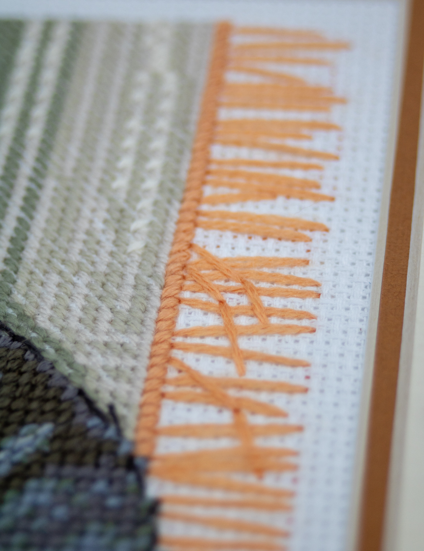 A completed cross-stitch of two cats resting on a rug, playing with orange yarn balls, stitched in warm brown, grey, and orange tones.