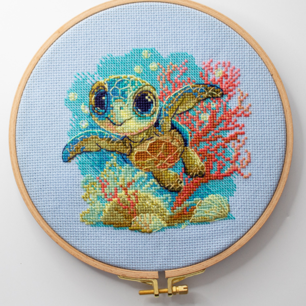 Cross-stitch kit showing a baby sea turtle underwater with coral, seashells, and a blue ocean background.