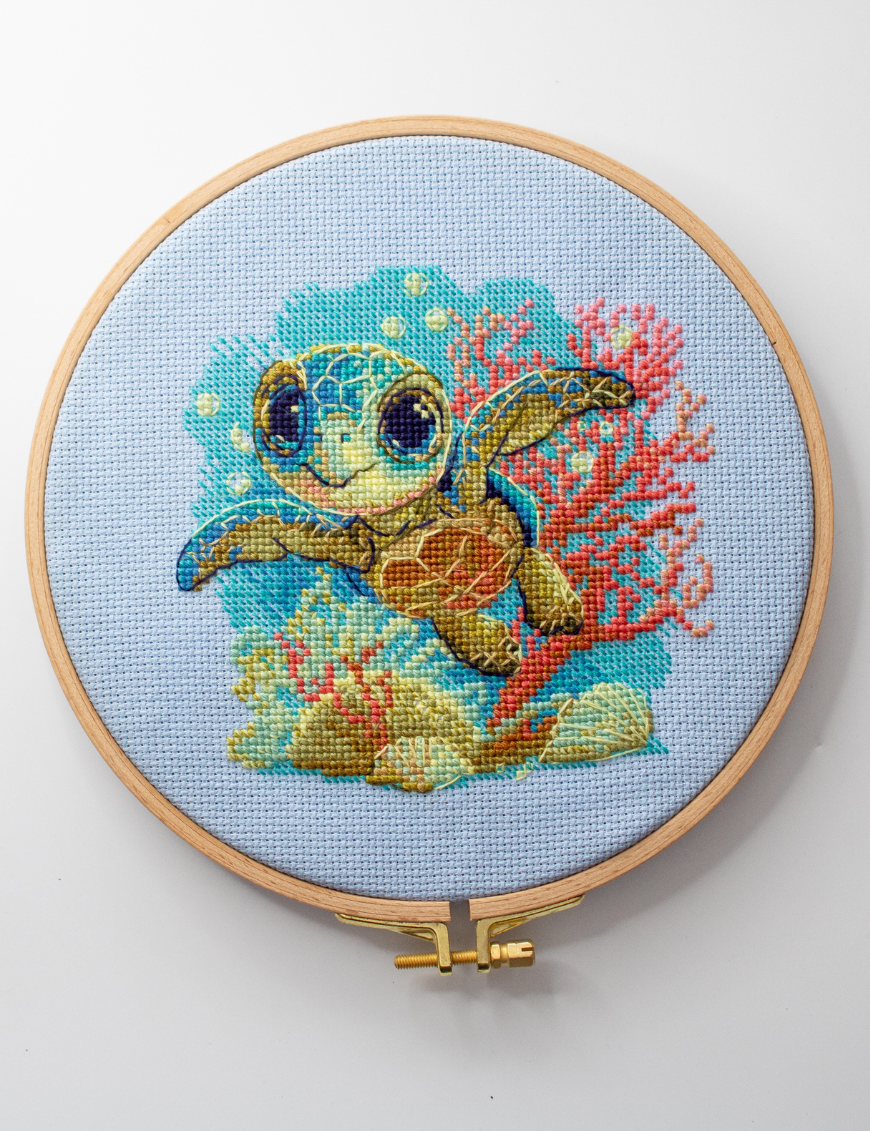 Cross-stitch kit showing a baby sea turtle underwater with coral, seashells, and a blue ocean background.