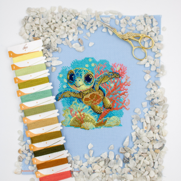 Cross-stitch kit showing a baby sea turtle underwater with coral, seashells, and a blue ocean background.