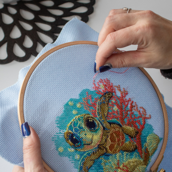 Cross-stitch kit showing a baby sea turtle underwater with coral, seashells, and a blue ocean background.