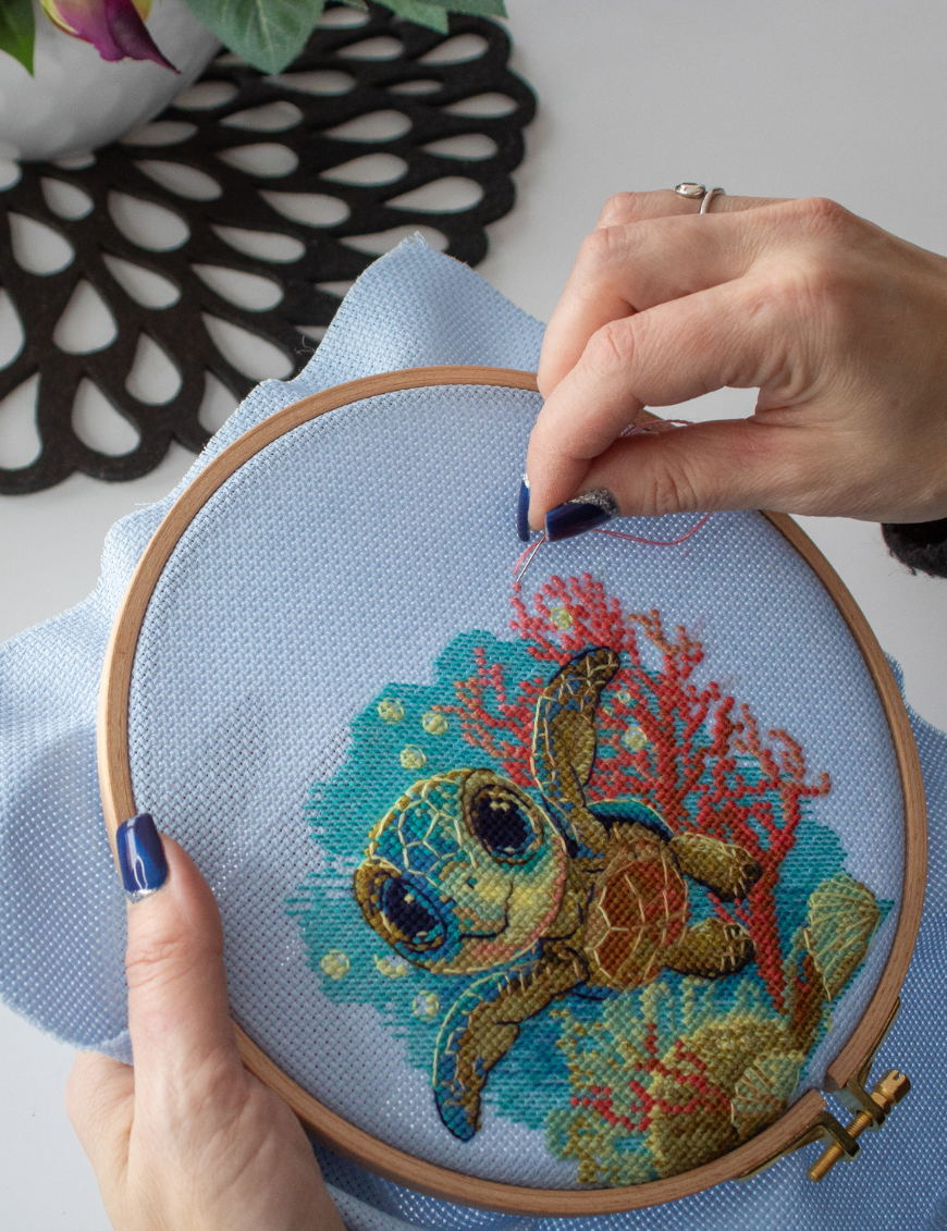 Cross-stitch kit showing a baby sea turtle underwater with coral, seashells, and a blue ocean background.