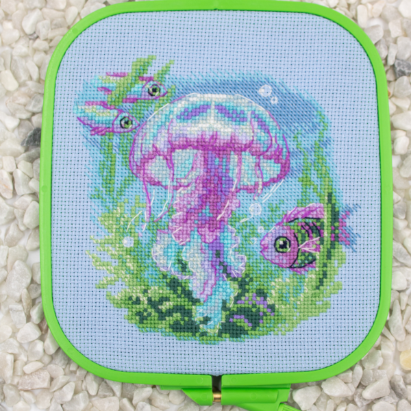 Baby Jellyfish cross stitch kit showing a pink and purple jellyfish underwater with green sea plants, bubbles, and three fish.