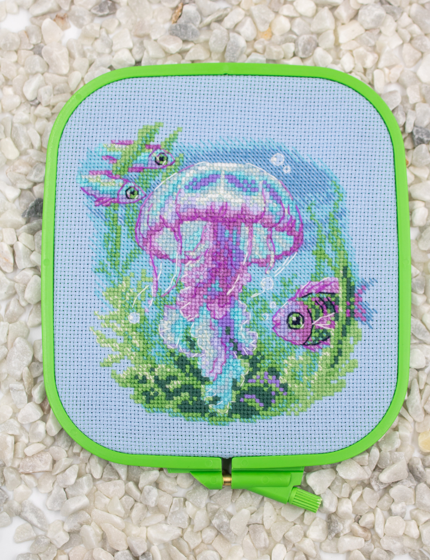 Baby Jellyfish cross stitch kit showing a pink and purple jellyfish underwater with green sea plants, bubbles, and three fish.