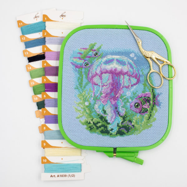 Baby Jellyfish cross stitch kit showing a pink and purple jellyfish underwater with green sea plants, bubbles, and three fish.