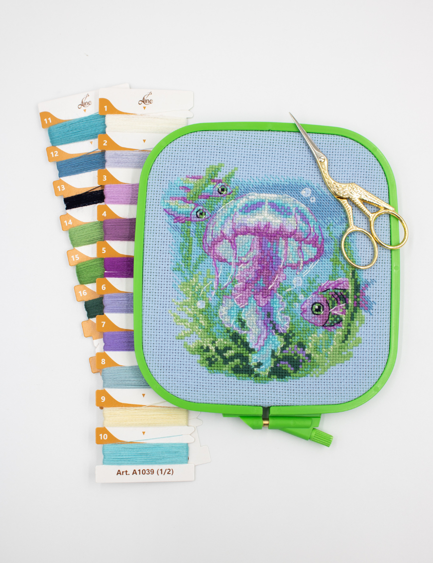 Baby Jellyfish cross stitch kit showing a pink and purple jellyfish underwater with green sea plants, bubbles, and three fish.