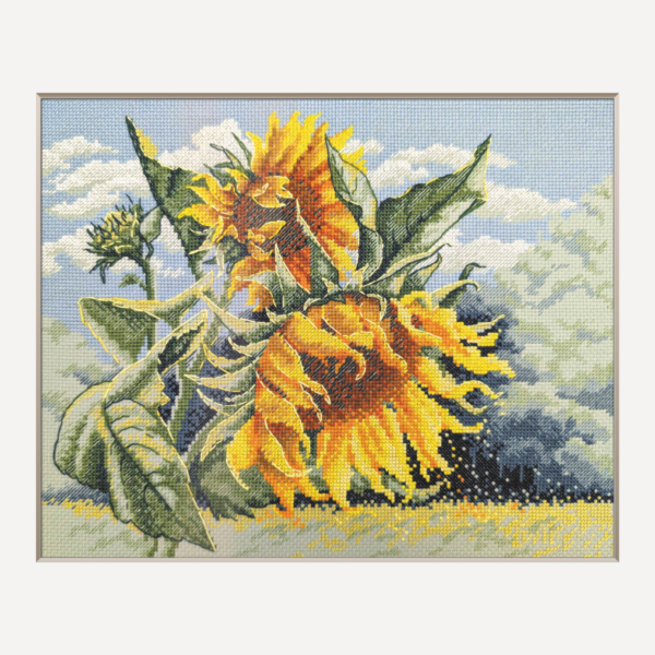 A completed cross-stitch of two large, vibrant yellow and orange sunflowers with textured green leaves, depicted as swaying in the wind against a light blue sky with fluffy white clouds.