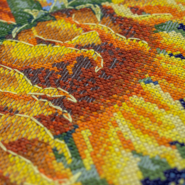 A completed cross-stitch of two large, vibrant yellow and orange sunflowers with textured green leaves, depicted as swaying in the wind against a light blue sky with fluffy white clouds.