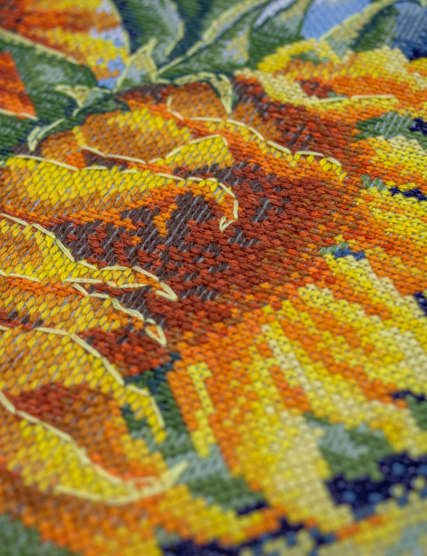 A completed cross-stitch of two large, vibrant yellow and orange sunflowers with textured green leaves, depicted as swaying in the wind against a light blue sky with fluffy white clouds.