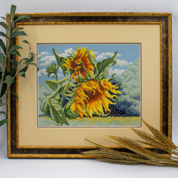 A completed cross-stitch of two large, vibrant yellow and orange sunflowers with textured green leaves, depicted as swaying in the wind against a light blue sky with fluffy white clouds.